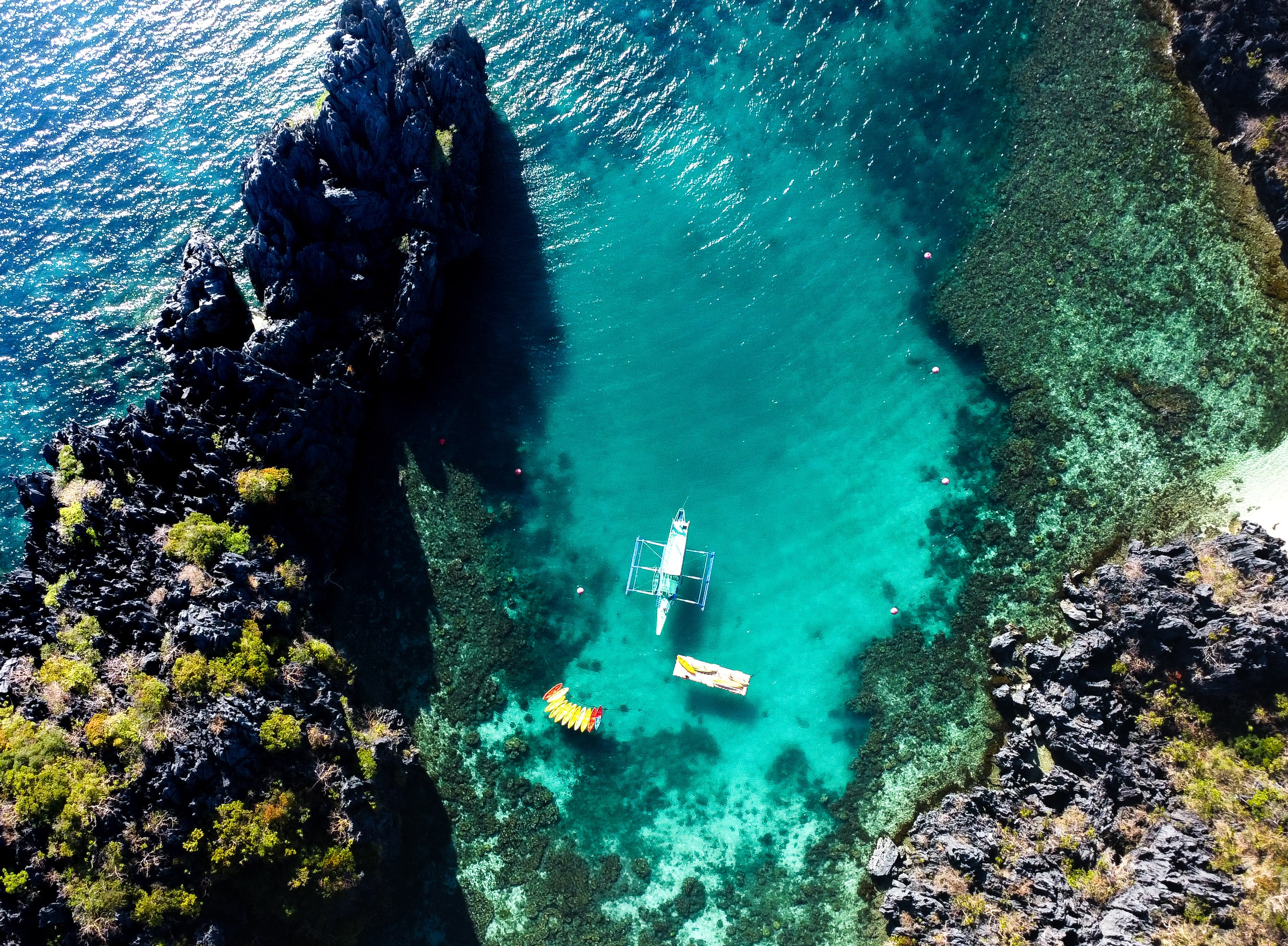 Looking down into a sheltered lagoon of clear turquoise water between black jagged limestone cliffs, where an outrigger boat, a small white boat and a yellow kayak float above visible reef.