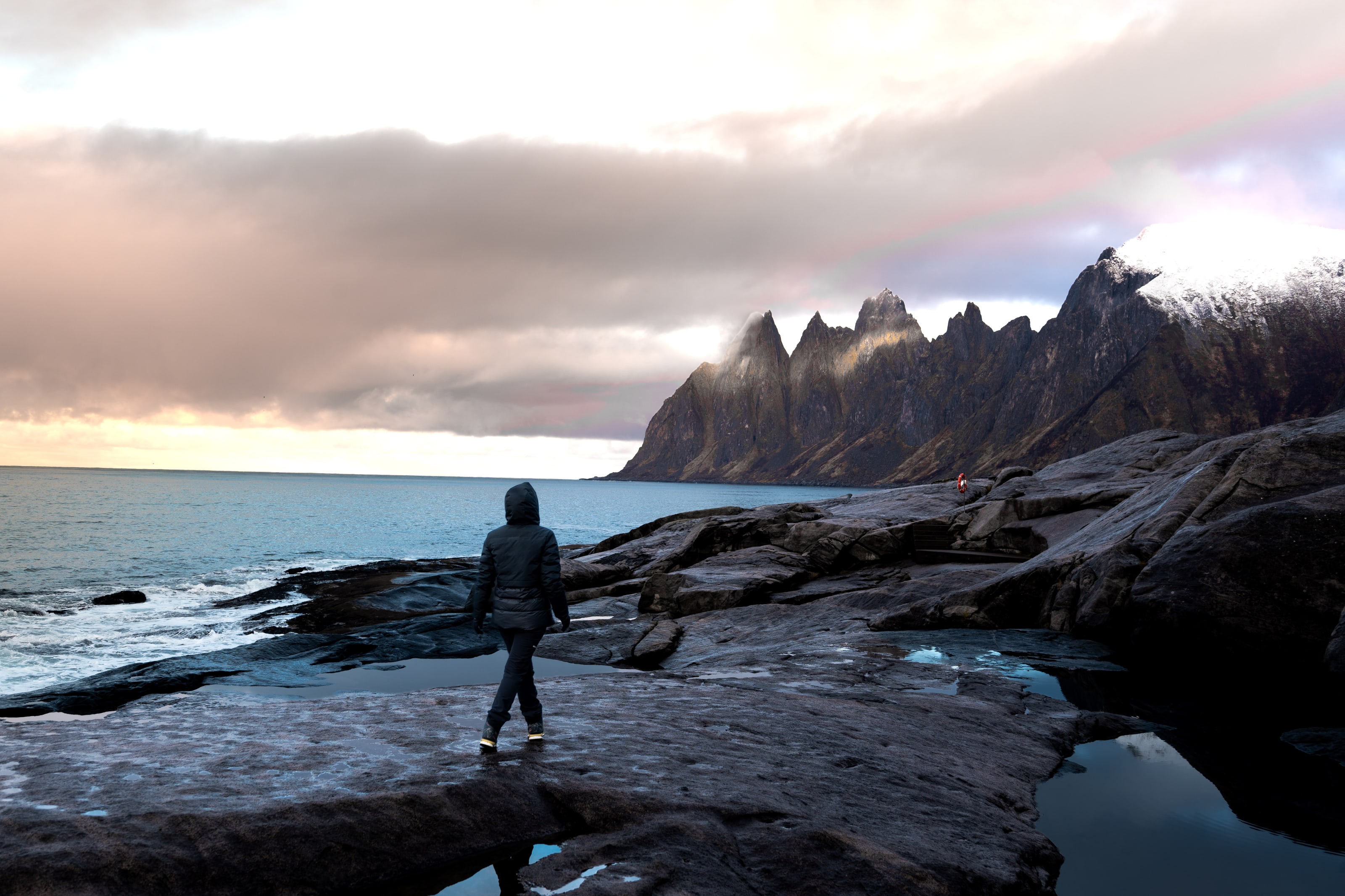 A lone figure in a dark hooded coat stands on a broad shelf of wet rock, facing a row of jagged mountain peaks across a grey sea under a low bank of sunset cloud.