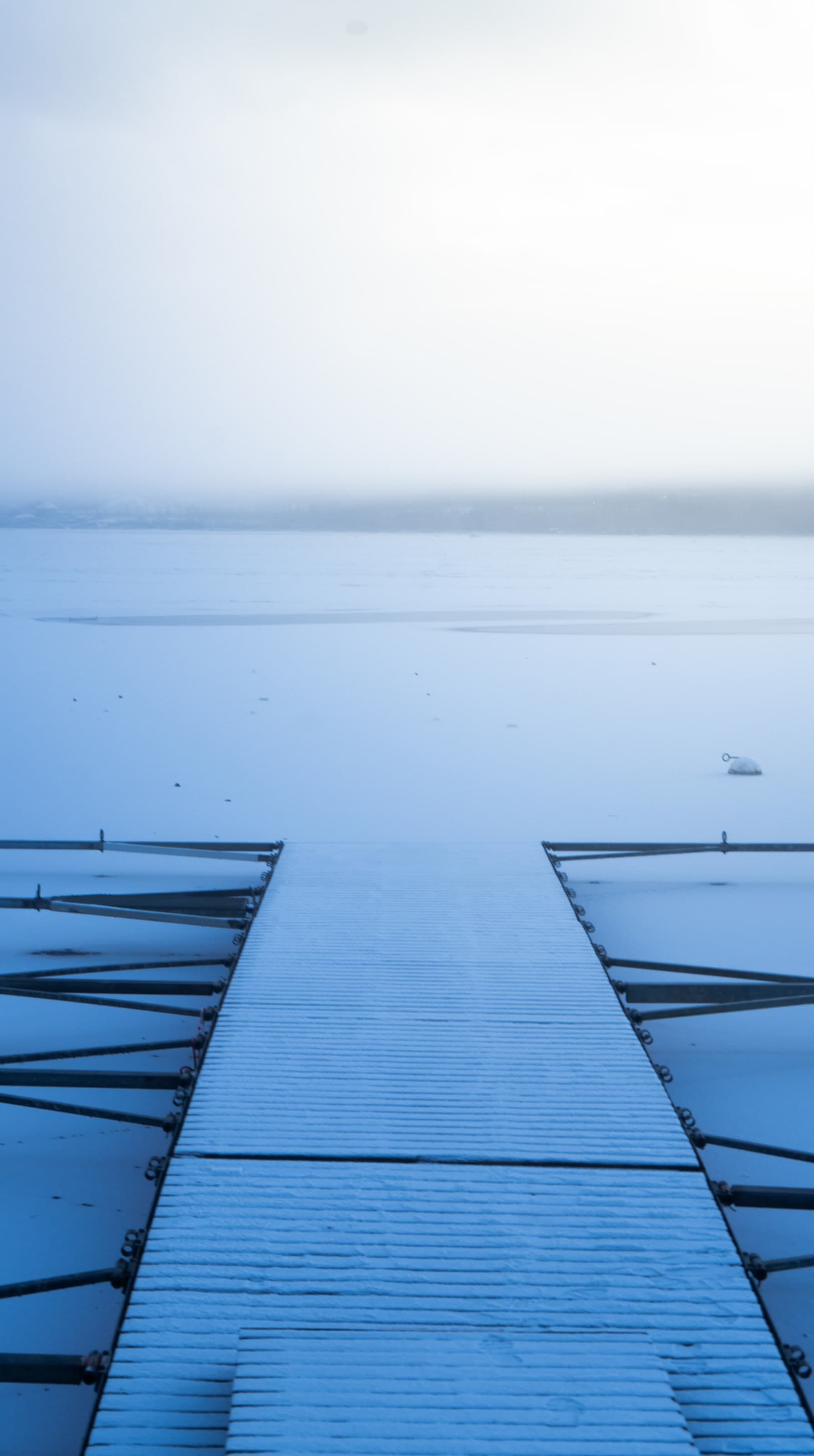 A narrow wooden jetty runs straight away from the viewer across a frozen, snow-dusted lake, its far end dissolving into white fog where the shoreline should be.