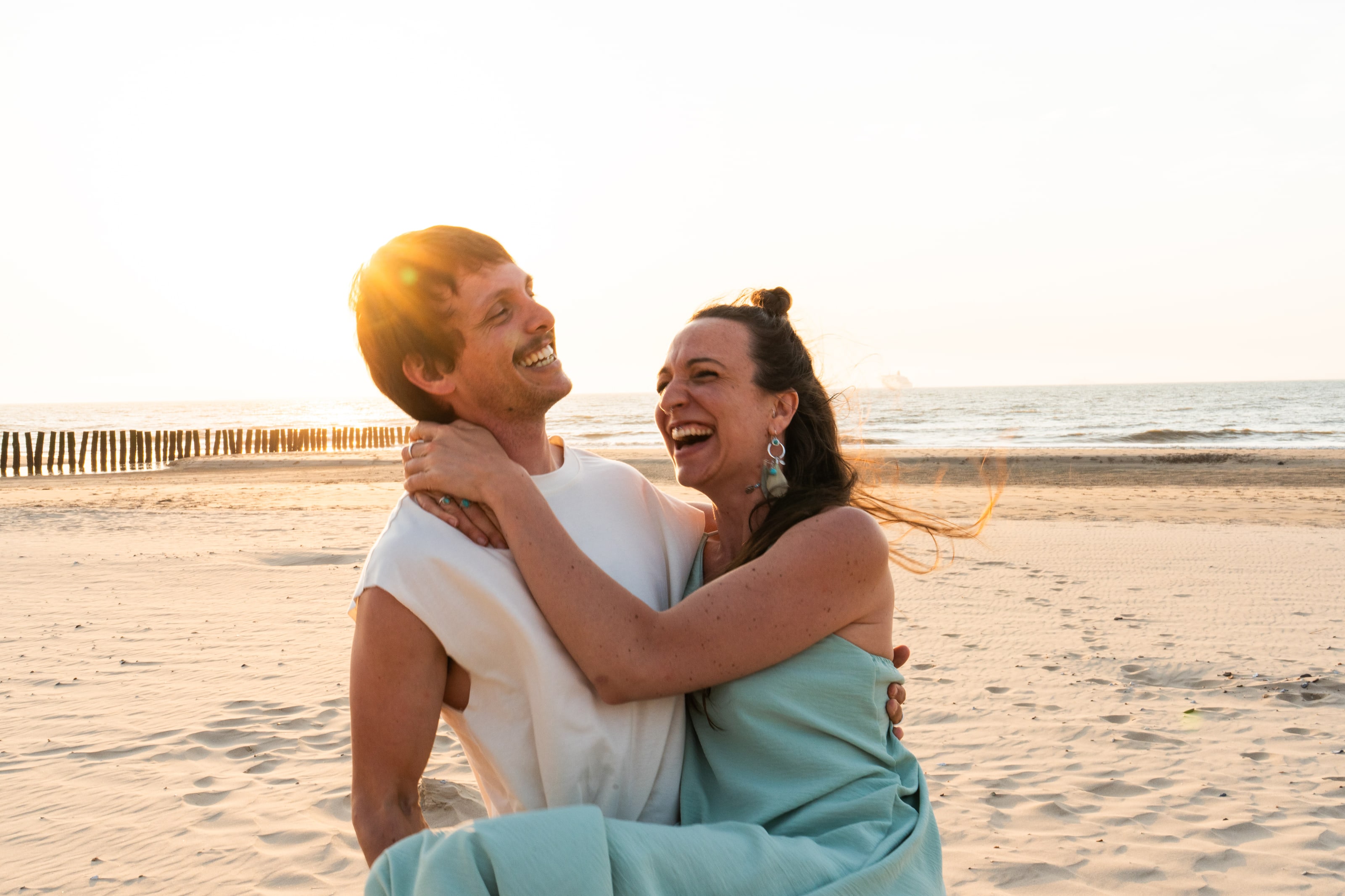 On a wide sandy beach at sunset, a woman with her arms around a man's neck laughs with her head thrown back, her hair blown across the frame, the low sun flaring behind him.