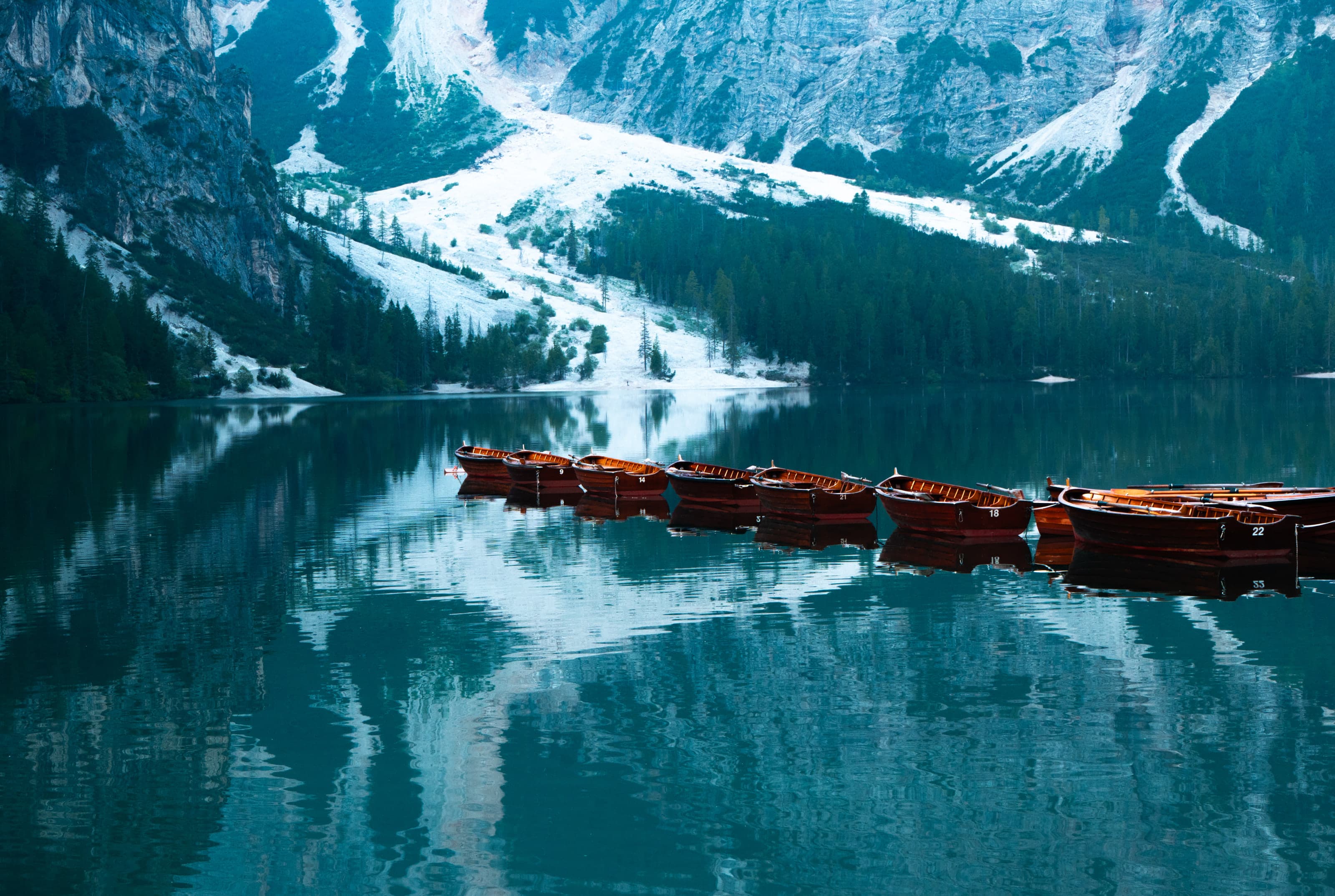 A row of varnished wooden rowing boats lies moored side by side on a still alpine lake of deep teal water, which is holding the reflection of a pale limestone face and dark conifer forest.