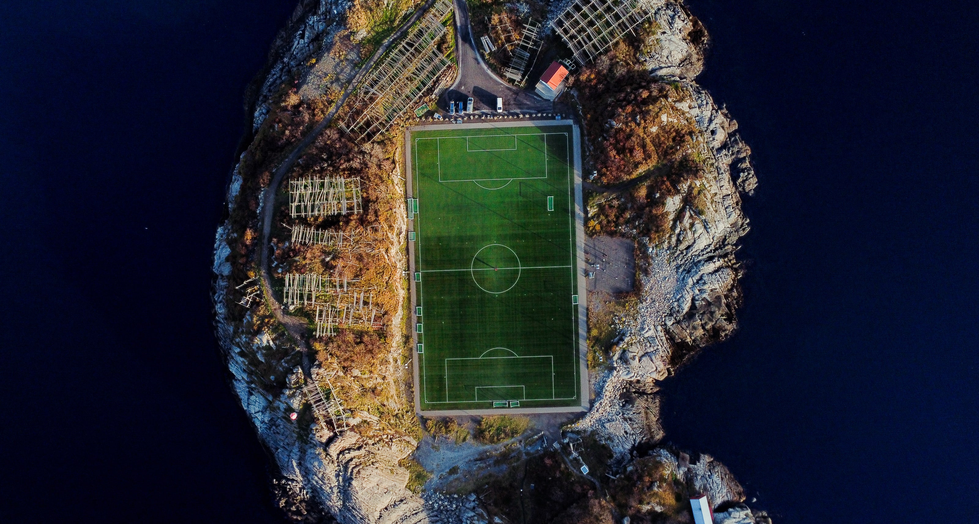Photographed from directly overhead, a full-size green football pitch fills a small rocky island, ringed by autumn scrub, timber drying racks and deep navy sea on every side.