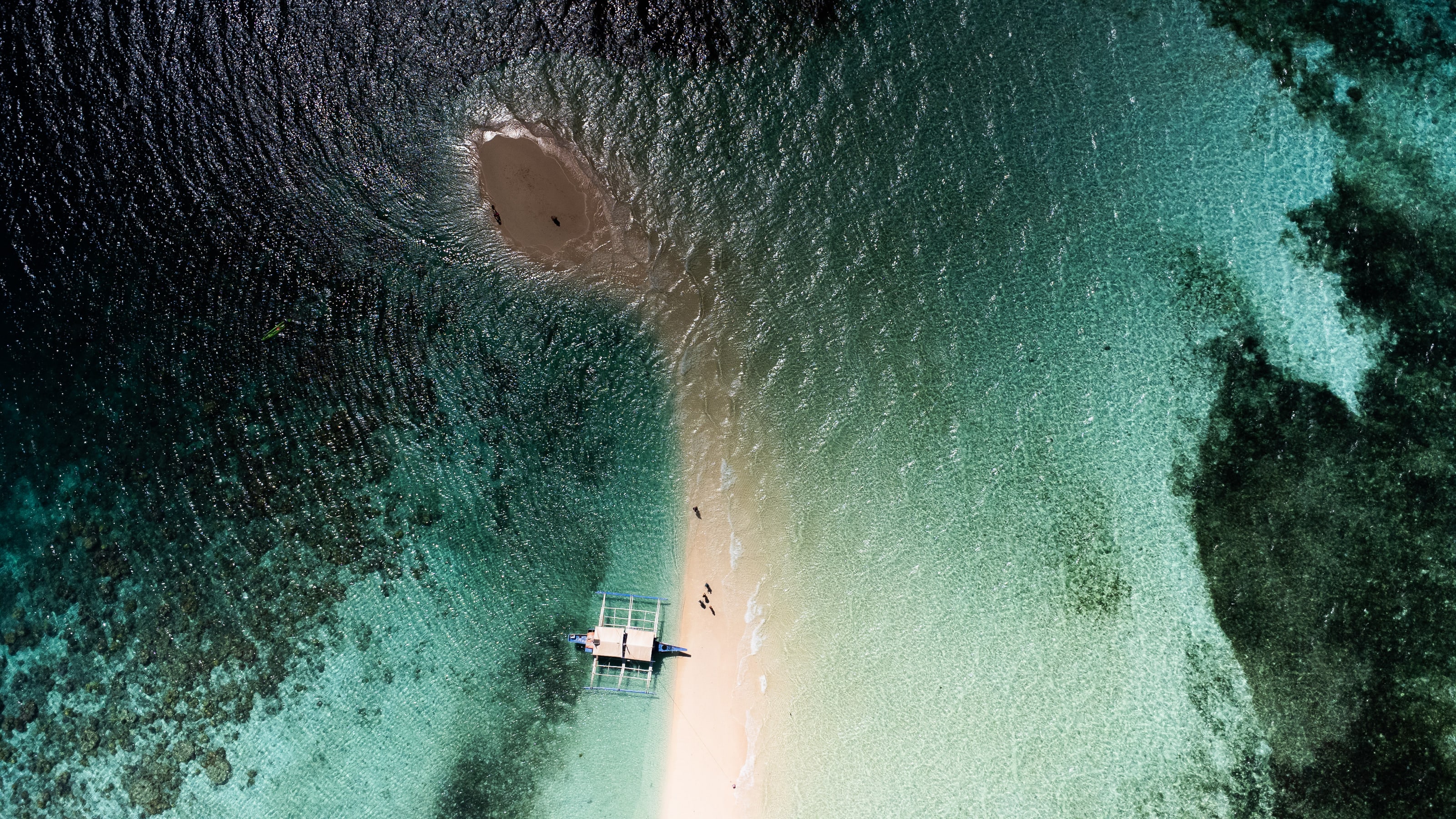 From directly above, a pale sand spit curves through shallow turquoise water between dark patches of reef, with a single white outrigger boat moored at its edge and a few tiny figures walking the sand.