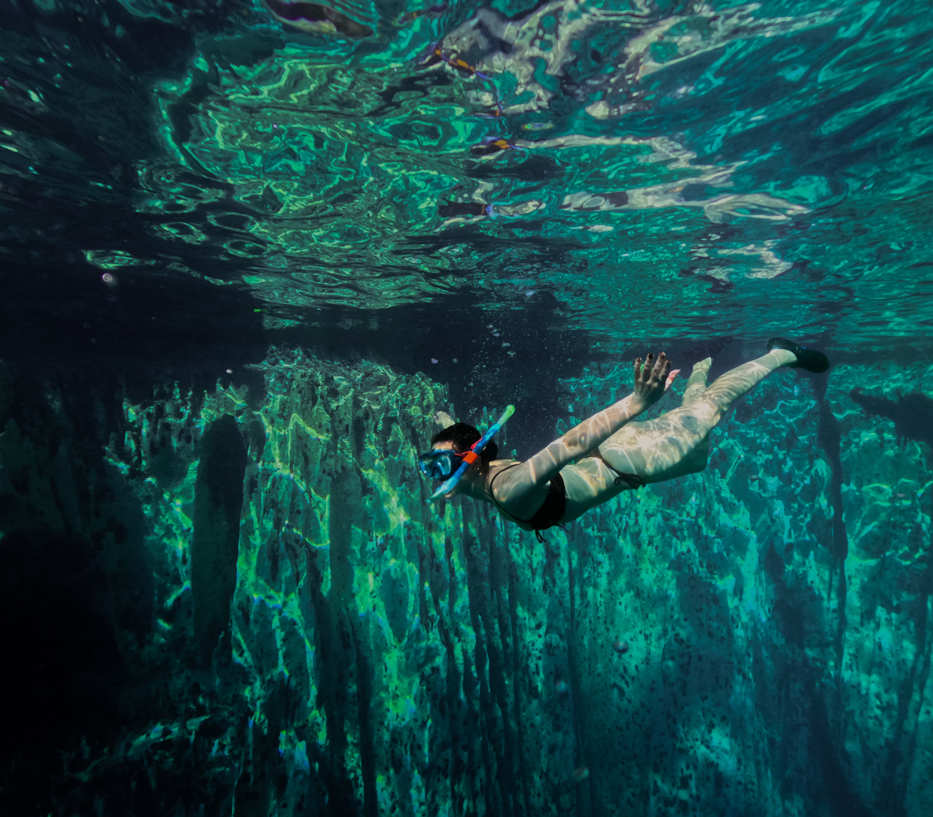 Photographed from underwater, a snorkeller in mask and fins glides just beneath the mirrored surface, above a fluted limestone wall that drops away into green darkness.