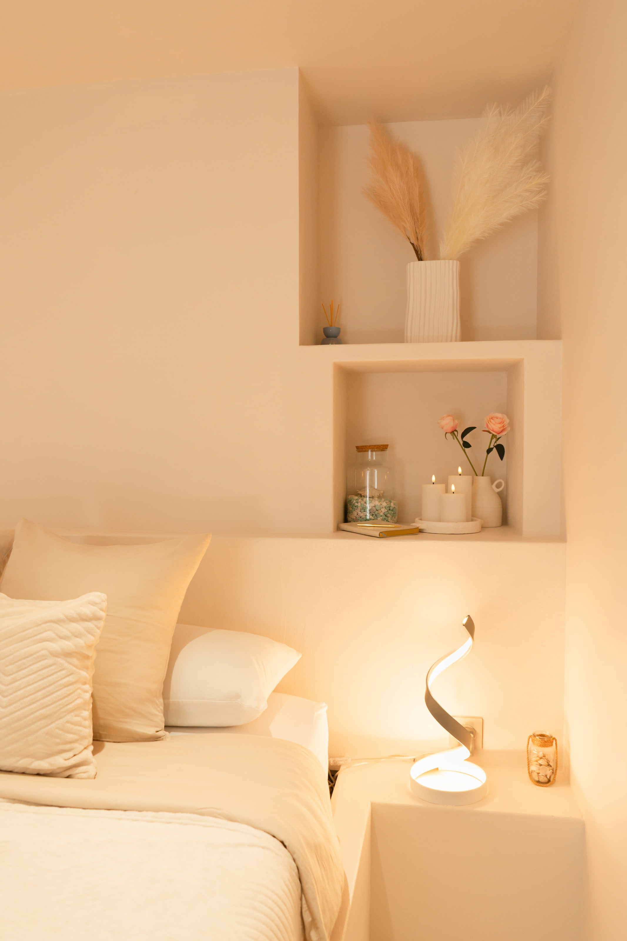 The corner of a bed in a cream plaster room, with two recessed niches above it — one holding pampas grass in a ribbed white vase, the other candles, two pink roses in a jug and a small book — and a curled sculptural lamp on the ledge below throwing warm light up the wall.