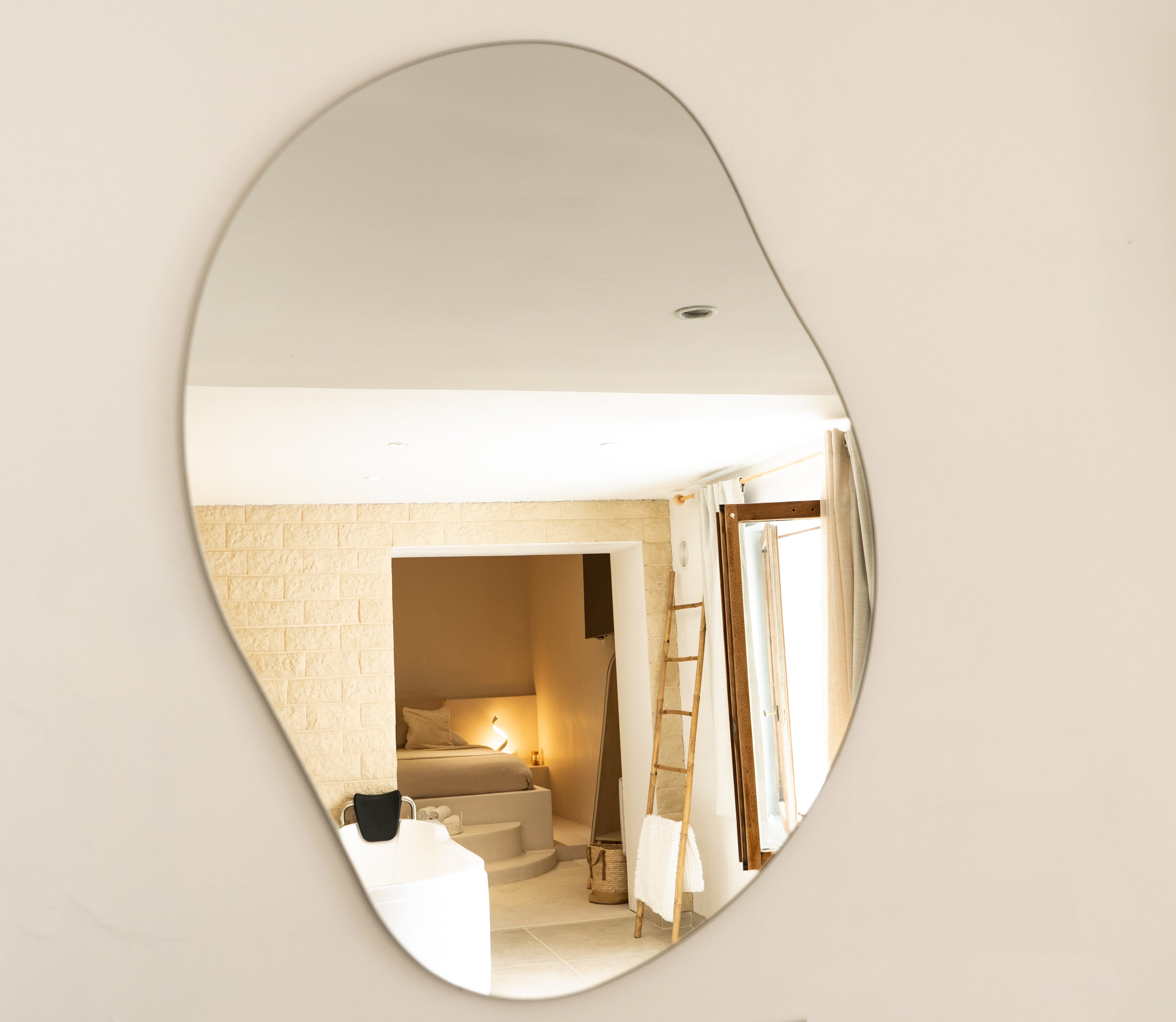 Un espejo sin marco, con forma de guijarro, cuelga solo en una pared de yeso desnuda y refleja la habitación que queda tras la cámara: un muro de bloques de piedra, una cama encajada en una alcoba con una lamparita encendida, una escalera de madera con una toalla blanca, una ventana abatible abierta y el borde de una bañera exenta.