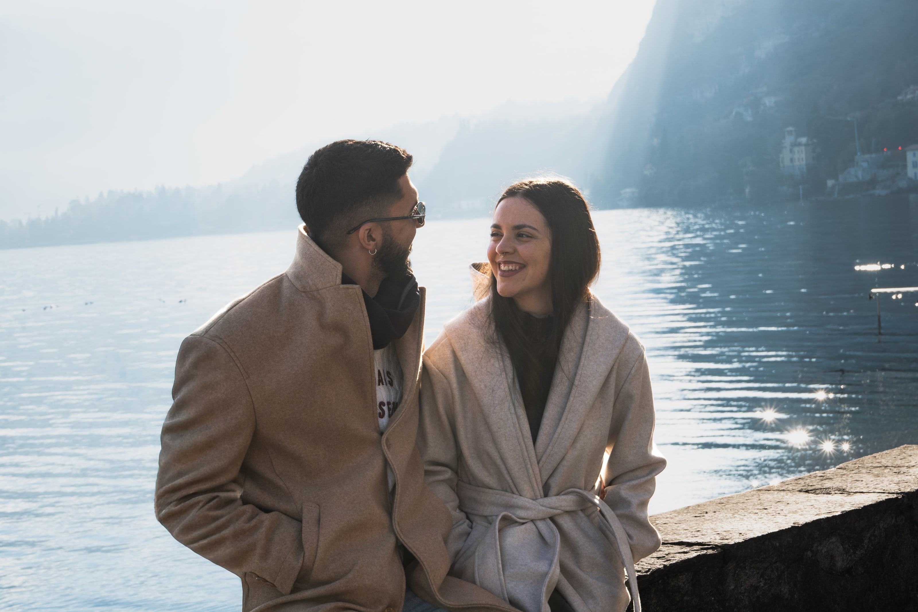 A couple in pale wool coats sit on a stone wall at the edge of a lake, turned toward each other mid-laugh, with low sun scattering in points of light across the water behind them.